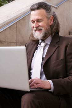 positive mature male entrepreneur in elegant suit siting on urban stairway and working online on laptop in city, concept of business and middle age man lifestyle