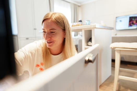 A woman with long, blonde hair is opening a white cabinet door in a modern kitchen. She is wearing a white shirt with a small orange design.