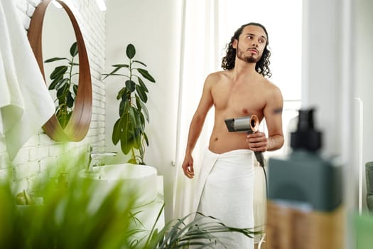 A man with curly hair styles himself in a bright room while using a hairdryer to enhance his look close up