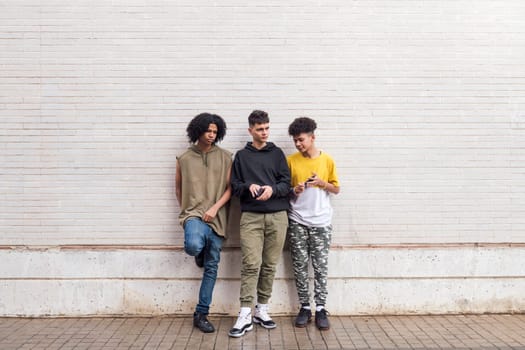 full body of three multiethnic teenager friends leaning against white brick wall, concept of friendship in youth and urban lifestyle, copyspace for text