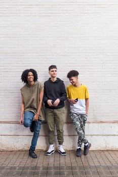 full body of three multiethnic teenager friends leaning against white brick wall, concept of friendship in youth and urban lifestyle, copyspace for text