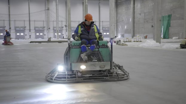 A skilled worker operates a concrete finishing machine to smooth a floor in a large industrial space. Clip