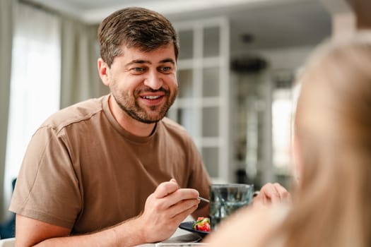A man with a beard sits at a table, smiling while holding a fork and eating. He is having a conversation with someone off-screen.