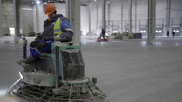 A skilled worker operates a concrete finishing machine to smooth a floor in a large industrial space. Clip