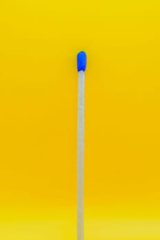 Close-up of a single wooden matchstick with a blue tip, isolated against a vibrant yellow background.