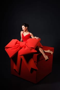 girl with big red bow, sitting on huge red gift box, on dark background, surprise, trend,