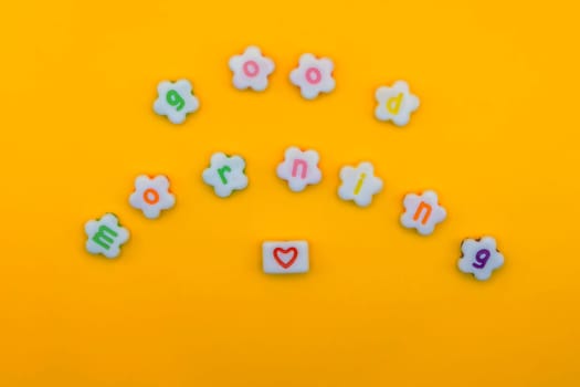 "Good morning" spelled with colorful flower-shaped letters and a red heart tile on a bright yellow background, cheerful and positive.