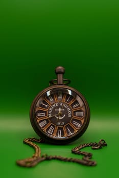 A vintage pocket watch featuring Roman numerals and "Marine Anchor 1912" design, displayed with a chain on a solid green backdrop.