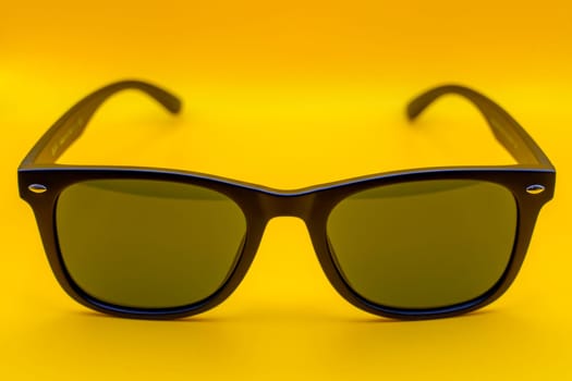 Minimalist close-up of black sunglasses with green-tinted lenses, centered against a vibrant yellow background.