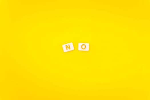 Minimalist arrangement of alphabet tiles spelling "No" with colorful letters on a bright yellow background, symbolizing negativity or refusal.
