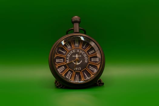 A close-up of a vintage pocket watch with Roman numerals and "Marine Anchor 1912" design, placed against a clean green background.