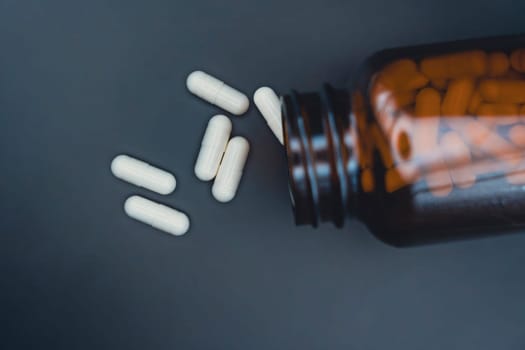 Top view of a brown glass bottle tipped over with white capsules scattered on a dark surface, representing healthcare, supplements, or medication.