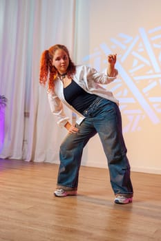 A young woman with red hair is dancing in a studio. She is wearing a white shirt and blue jeans. The floor is made of wood. She is standing with her right leg bent and her left leg straight.