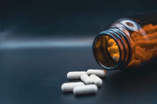 A brown glass bottle tipped over with white capsules spilling onto a dark background, representing healthcare, medication, or dietary supplements.
