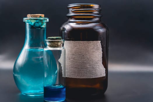 Close-up of three glass bottles, one brown with a torn label and two corked blue bottles containing liquid, placed against a dark background.
