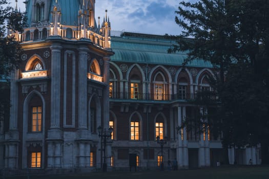 Detailed view of a palace facade with Gothic arches and warm illuminated windows, surrounded by trees during twilight.