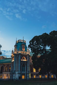 A grand palace tower with Gothic details and illuminated windows, surrounded by trees, captured under a clear evening sky.