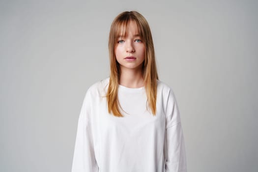 Young woman with long hair in a white sweatshirt standing against a light gray background close up