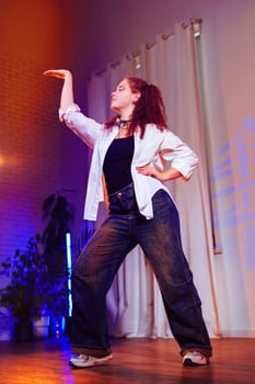 A young woman with red hair dances in a studio. She is wearing a white shirt, blue jeans, and white sneakers. The studio is lit with colorful lights, and there are white curtains in the background.