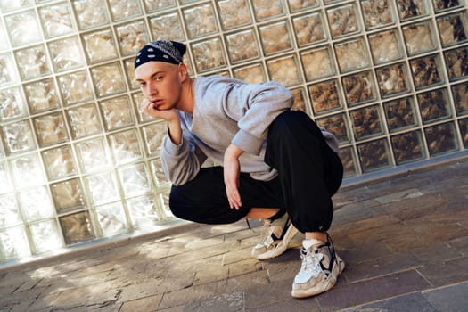 A man wearing a gray sweatshirt and a black bandana sits in a squat position in front of a wall made of clear glass blocks.