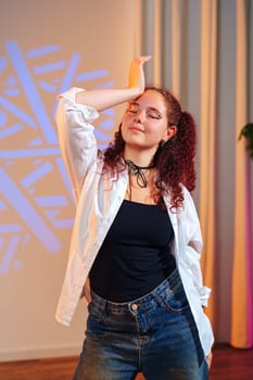 A young woman with red curly hair is wearing a white button-down shirt over a black tank top and blue jeans. She has her hand on her head and is looking towards the camera with her eyes closed.