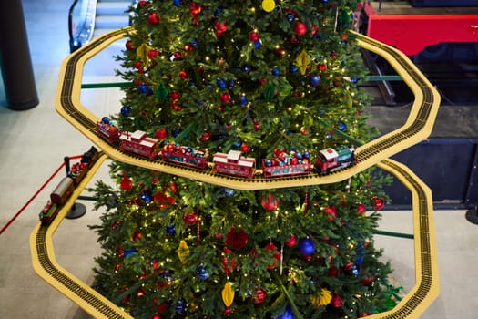 A colorful Christmas tree, adorned with ornaments, features a toy train set circling around its base