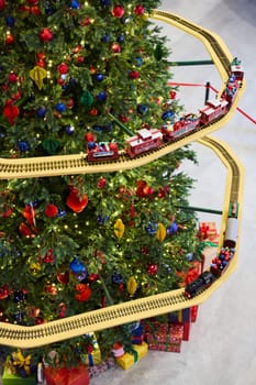 A beautifully vibrant Christmas tree is adorned with colorful, shiny ornaments and has a festive train track around it