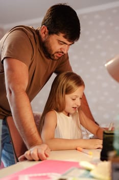 A father stands over his young daughter as she sits at a table working on her homework. He is looking down at her, watching her as she uses a pencil and paper.