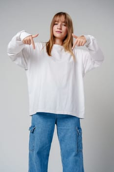 A young girl in a white long-sleeve shirt smiles while gesturing downwards in a light-colored room close up