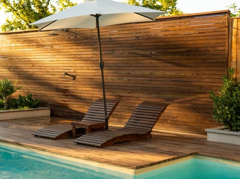 Sunlit poolside area with sleek wooden deck, comfortable loungers, and a large umbrella for shade