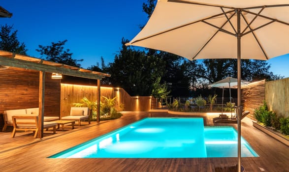 Backyard swimming pool with wooden deck and large umbrella in cozy ambiance. Luxury outdoor relaxation area with soft evening lighting