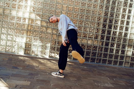 A young man in a gray sweatshirt and black pants performs a dance move in front of a wall made of glass blocks. His right foot is raised high in the air, demonstrating his flexibility and athleticism.
