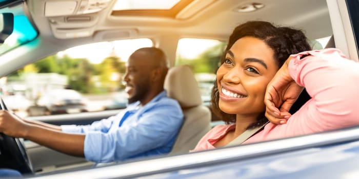 Enjoying Travel Concept. Smiling handsome African American man driving a car, selective focus on happy beautiful young woman sitting on the front passenger seat and looking out of window