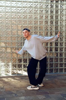 A man stands in front of a wall of glass blocks. He is wearing a grey sweatshirt, black pants, and white sneakers. He is looking at the camera and has his right arm outstretched.