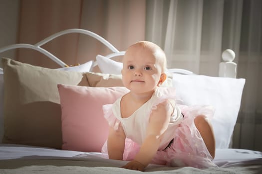 A baby in a pink tutu crawls on a bed surrounded by soft pillows, exploring the cozy space.