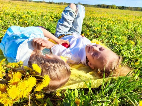 Two children are relaxing on a yellow blanket in a vibrant dandelion field, smiling and enjoying a sunny day outdoors.