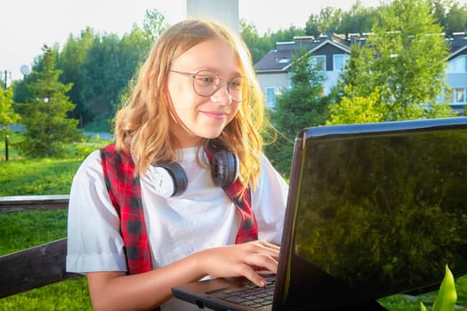 In a bright outdoor area, a young girl happily works on her laptop while enjoying the lush surroundings in the background.