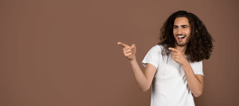 A young Caucasian man with long, curly hair joyfully poses while pointing at copy space. His carefree expression reflects a vibrant and playful attitude toward life.