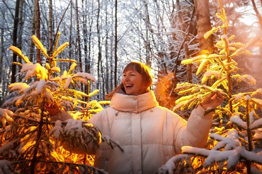 A middle-aged woman joyfully walks through a snow-laden forest, surrounded by beautifully frosted trees, celebrating winter and the holiday spirit.