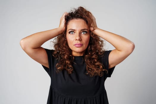 Smiling woman with curly hair poses confidently against a plain background while holding her hands behind her head indoors
