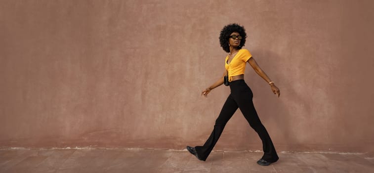 A trendsetter strolls effortlessly along a brown wall, showcasing a bright yellow top paired with black flared pants. The confident posture and cool sunglasses enhance their fashion statement.