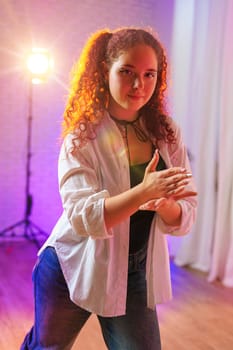 A young woman with long, curly red hair, wearing a white button-down shirt and blue jeans, practices a dance move in a studio with purple lighting.