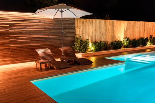 Luxury swimming pool with a wooden deck and stylish sun loungers under a night sky. Concept of a modern and peaceful outdoor space for relaxation and leisure