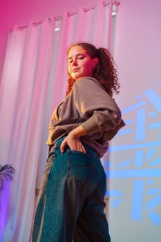 A young woman with curly brown hair stands facing the camera, wearing a brown sweatshirt and blue jeans. She has one hand in her pocket, and her other arm is bent at the elbow.