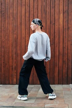 A person wearing a grey sweatshirt, black pants, and a black bandana walks past a wooden wall with vertical planks. The person is walking away from the camera, and their right foot is in the air.