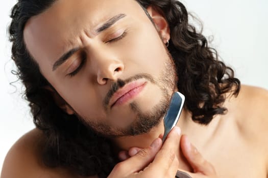 Man with curly hair grooming his beard carefully using a straight razor against a plain white background close up