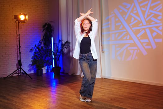 A woman dances in a studio with a white shirt and blue jeans. She is in front of a white wall with a blue projector light shining on it. The room is lit with other blue and yellow lights.