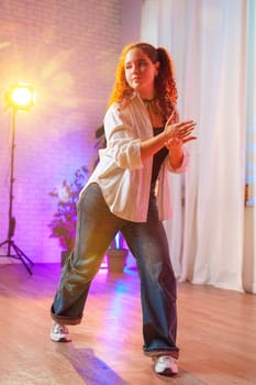 A young woman with curly red hair dances in a studio, wearing a white shirt, blue jeans, and white sneakers. She has her arms raised, and she is looking to the side.