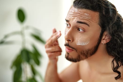 A young man applies skincare cream while looking in a round mirror in a bright bathroom close up