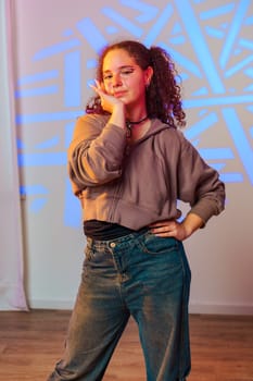 A young woman with curly brown hair is standing in a dance studio. She is wearing a brown cropped hoodie, a black top, and blue jeans. She has one hand on her chin and the other on her hip.